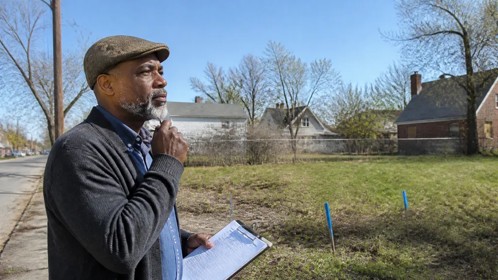 A landowner stands on the street with paperwork, looking over his vacant lot with survey stakes in the grass.