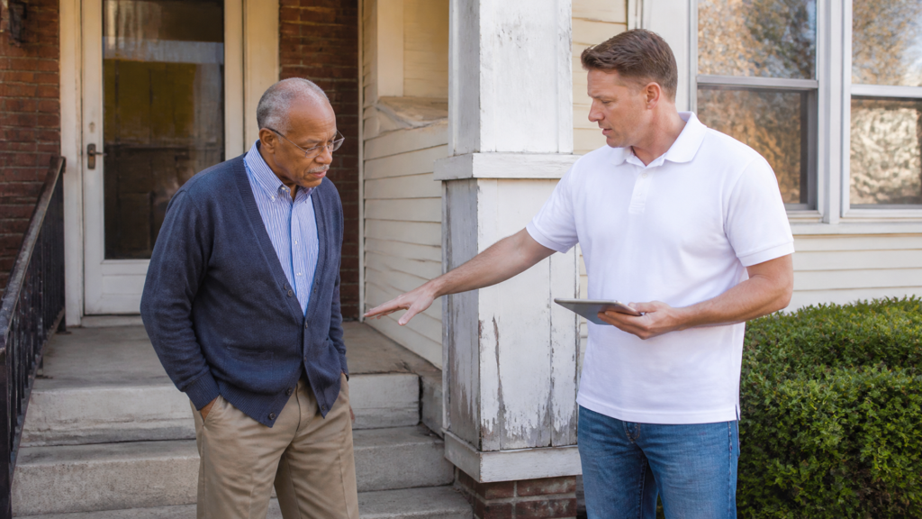 A buyer points out peeling siding beside the porch while a homeowner looks on, tablet in the buyer's hand.
